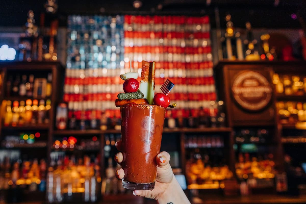 Bloody Mary cocktail in a glass being held up by a hand in front of Bub City's beer can American flag