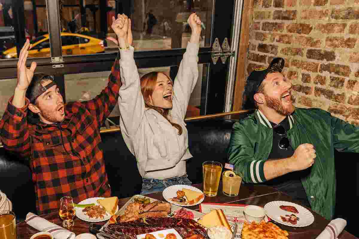 Three people sitting at a restaurant table cheering and watching a game on TV