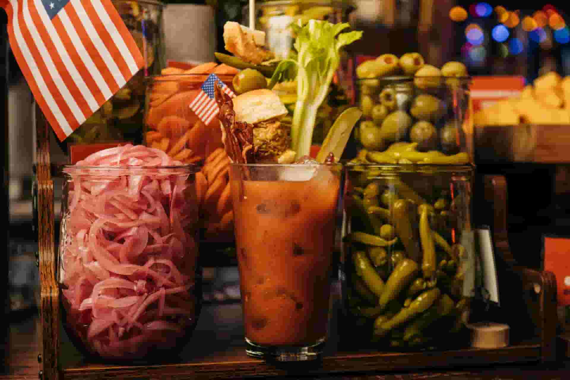 Bloody Mary cocktail in a clear pint glass sitting on a wooden shelf surrounded by mason jars full of garnishes